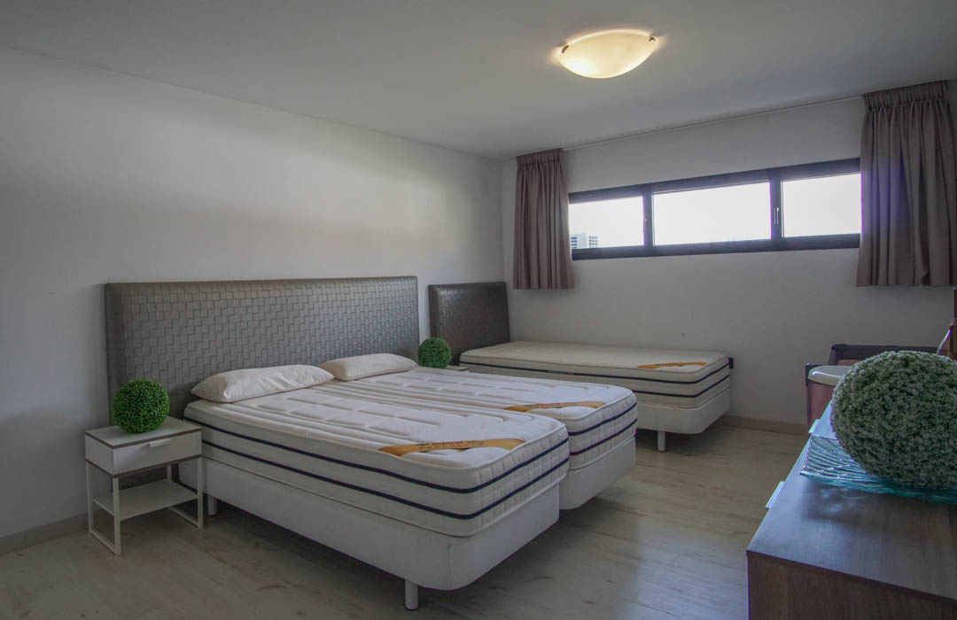 Slaapkamer met twee samengevoegde eenpersoonsbedden, grijze gestructureerde hoofdeinden, een derde eenpersoonsbed achterin, lichte houten vloer en horizontale ramen.