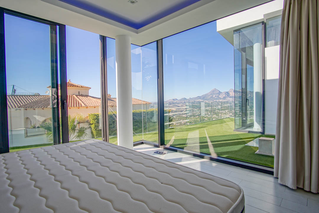 Slaapkamer met uitgebreide glazen hoekwanden, met panoramisch uitzicht op verre bergen, een vallei en naburige daken. Voorzien van een witte structurele pilaar en toegang tot kunstgras.