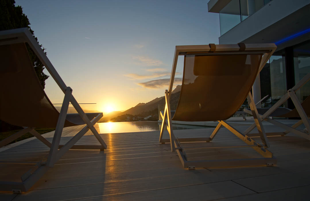 Twee moderne ligstoelen op een licht terras kijken uit op een overloopzwembad, dat de gouden zonsondergang over de bergen weerspiegelt.
