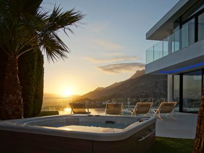 Buitenjacuzzi met houtlook, op groen gras, voor een moderne villa met glazen balkons. Uitzicht op bergen en zee tijdens zonsondergang.