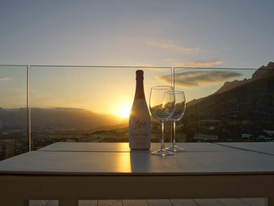 Buitentafel op terras met glazen balustrade, met fles rosé cava en twee glazen. Zonsondergang over bergen en vallei, met warme reflecties van het avondlicht.