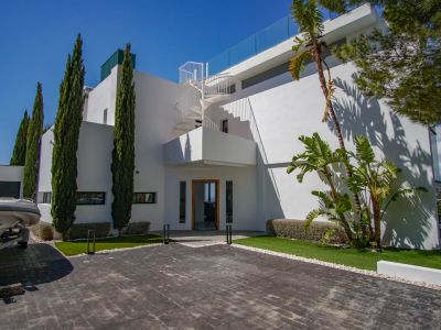 De moderne witte villa-exterieur omvat een geplaveide oprit, hoge cipressen en een opvallende wenteltrap naar het boventerras met glazen balustrade.
