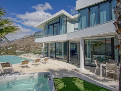 De moderne witte villa-exterieur omvat een overloopzwembad, aparte jacuzzi en een ruim terras met ligstoelen, omringd door berggezichten onder een heldere hemel.