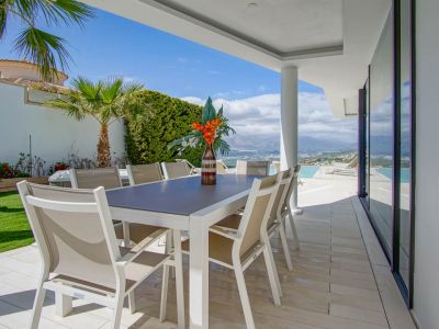 Dit overdekte buitenterras heeft een moderne witte tafel met acht beige stoelen op lichte tegels. Het biedt een weids uitzicht op de bergen en de zee, grenzend aan het overloopzwembad.