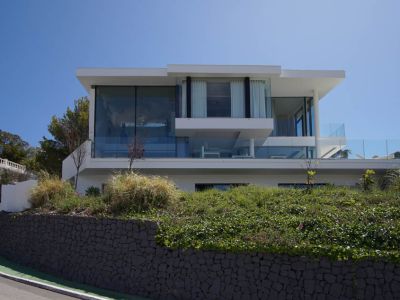 Exterieur van een moderne villa met drie verdiepingen, voorzien van uitgebreide glazen gevels en balustrades, verheven boven een stenen muur en vegetatie langs een bochtige weg.