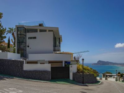 Exterieur van een moderne witte villa met glazen lift en dakterrasreling. Stenen keermuren en een automatische poort omlijsten het panoramische uitzicht op zee.