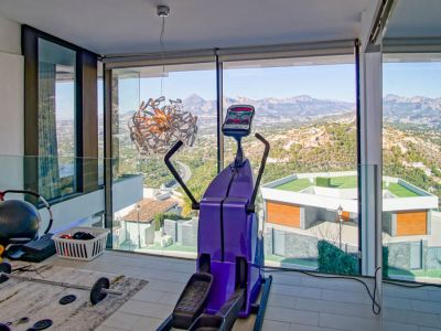 Fitnessruimte met kamerhoge ramen, met panoramisch uitzicht op bergen en vallei. Voorzien van een crosstrainer en gewichten, aangevuld met een glazen balustrade.