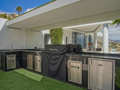 L-vormige buitenkeuken met zwart granieten werkbladen, dubbele spoelbakken, een overdekte BBQ en geïntegreerde RVS-apparatuur, met weerspiegelde zwembadzichten.