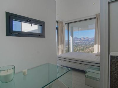 Lichte slaapkamer met bed voor kamerhoge ramen, met panoramisch uitzicht op bergen en vallei. Een glazen console en een horizontaal raam met donker kozijn zijn ook zichtbaar.
