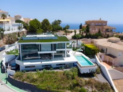 Luchtfoto van de moderne witte designvilla met groen dak, overloopzwembad en aparte jacuzzi, gelegen in een omgeving met zeezicht.