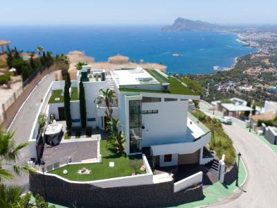 Luchtfoto van een moderne witte villa met architectuur op meerdere niveaus, groene platte daken en een externe glazen lift. Het pand omvat een privé-oprit en biedt panoramisch zee- en kustzicht.