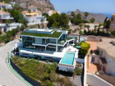 Luchtfoto van een moderne witte villa met overloopzwembad, glazen gevels, een groen dak en aparte jacuzzi, met uitzicht op bergen en zee.