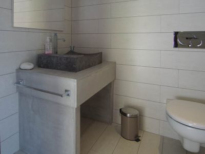 Modern gastentoilet met stenen waskom op betonnen wastafelmeubel en lichte tegels. Wandhangend toilet met chromen spoelplaat.