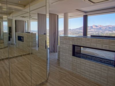 Modern interieur met een transparante gashaard op kiezelbasis, ingebouwd in een getextureerde muur. Spiegelpanelen reflecteren de kamer en het weidse berg- en zeezicht door kamerhoge ramen.