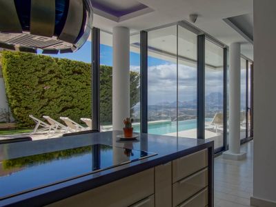 Modern kookeiland met donker, reflecterend werkblad en geïntegreerde kookplaat. Grote glazen deuren bieden uitzicht op de infinity pool en panoramische berg- en dalgezichten.