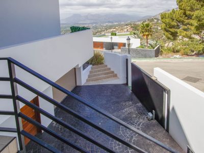 Moderne buitentrap met zwarte balustrades daalt af naar een donkergrijze, gestempelde oprit, voorzien van een automatische poort, garage-ingang en panoramisch bergzicht.