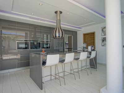 Moderne keuken met hoogglans grijze kasten, geïntegreerde ovens en een centraal eiland met zitplaatsen voor vier. Inbouwspots en paarse LED-stripverlichting in het plafond.
