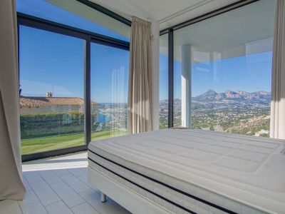 Moderne slaapkamer met groot bed en uitgestrekte ramen. Het biedt panoramisch uitzicht op de verre zee, een groen gazon en ruige bergen onder een heldere hemel.
