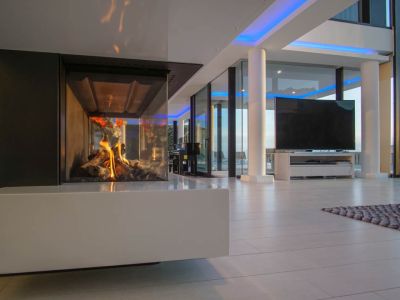 Moderne woonruimte met glazen gashaard en witte basis. Grote ramen en blauwe accentverlichting complementeren het ontwerp.