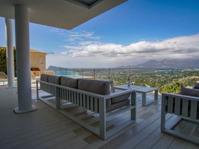 Ruim overdekt terras met lichte vlonder en modern wit loungemeubilair, dat via een glazen balustrade een panoramisch uitzicht op zee, kust en bergen omlijst.