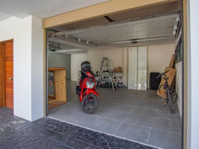 Ruime garage met grijze tegelvloer, een deels geopende sectionaaldeur en een opvallende houten toegangsdeur links. Binnen zijn een rode scooter en fiets zichtbaar.