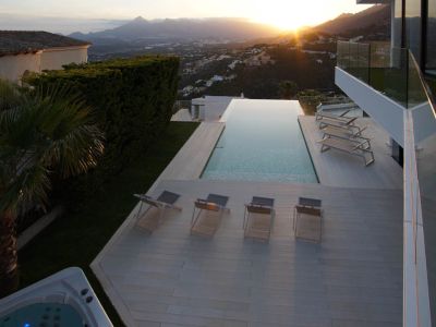 Ruime infinity pool met lichtgekleurd terras, diverse ligbedden en aparte jacuzzi, omkaderd door een panoramisch berglandschap bij zonsondergang.