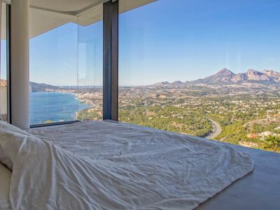 Slaapkamer met kamerhoge glazen ramen, die panoramisch uitzicht bieden op de Middellandse Zee, kustplaats en bergen. Een laag bed met wit beddengoed kijkt uit op het weidse panorama.
