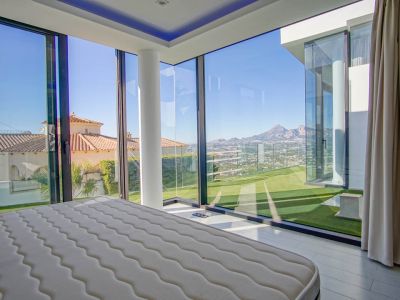 Slaapkamer met uitgebreide glazen hoekwanden, met panoramisch uitzicht op verre bergen, een vallei en naburige daken. Voorzien van een witte structurele pilaar en toegang tot kunstgras.