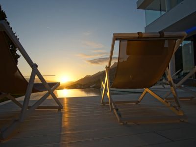 Twee moderne ligstoelen op een licht terras kijken uit op een overloopzwembad, dat de gouden zonsondergang over de bergen weerspiegelt.