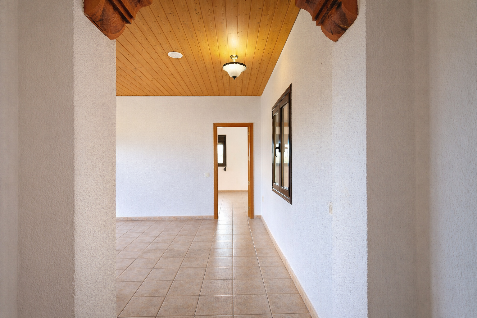 Binnenhal met lichte tegelvloer en gestructureerde witte muren. Houten plafond met centrale verlichting en gesneden decoratieve boogdetails.