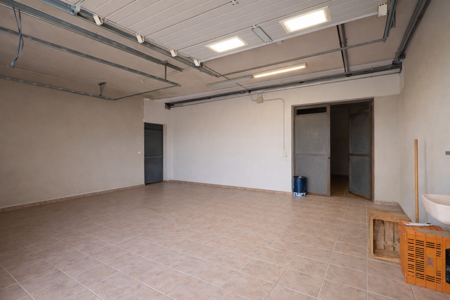 Ruime garage of technische ruimte met lichte tegelvloer, industriële plafondverlichting en meerdere toegangspunten.