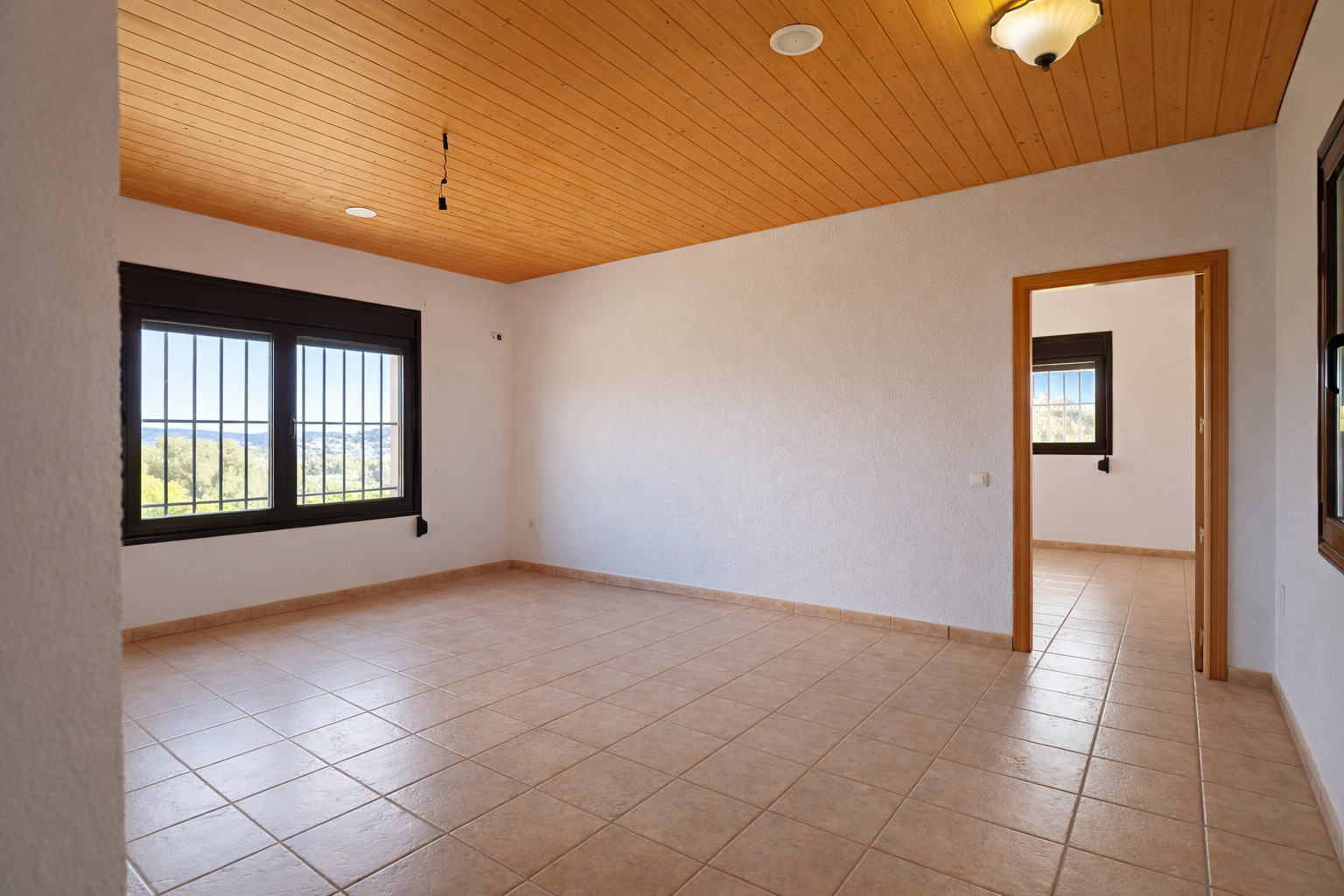 Ruime kamer met lichte tegelvloer, witte muren en een warm houten plafond. De ramen met tralies bieden een weids uitzicht op de vallei en veel natuurlijk licht.