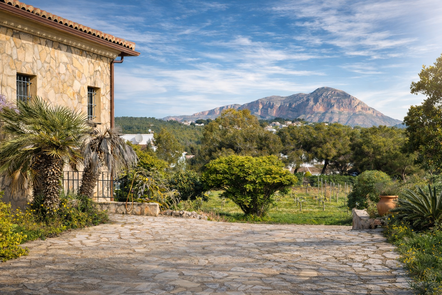Rustieke stenen gevel van de finca met palmbomen, geplaveide oprit en uitzicht op de Montgó, omringd door wijngaarden en mediterrane vegetatie.