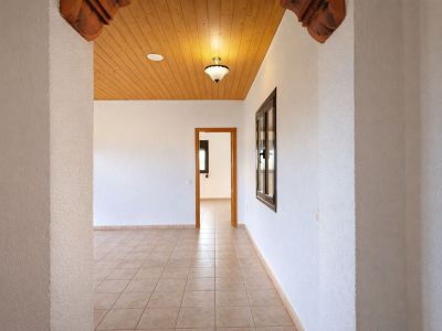 Binnenhal met lichte tegelvloer en gestructureerde witte muren. Houten plafond met centrale verlichting en gesneden decoratieve boogdetails.