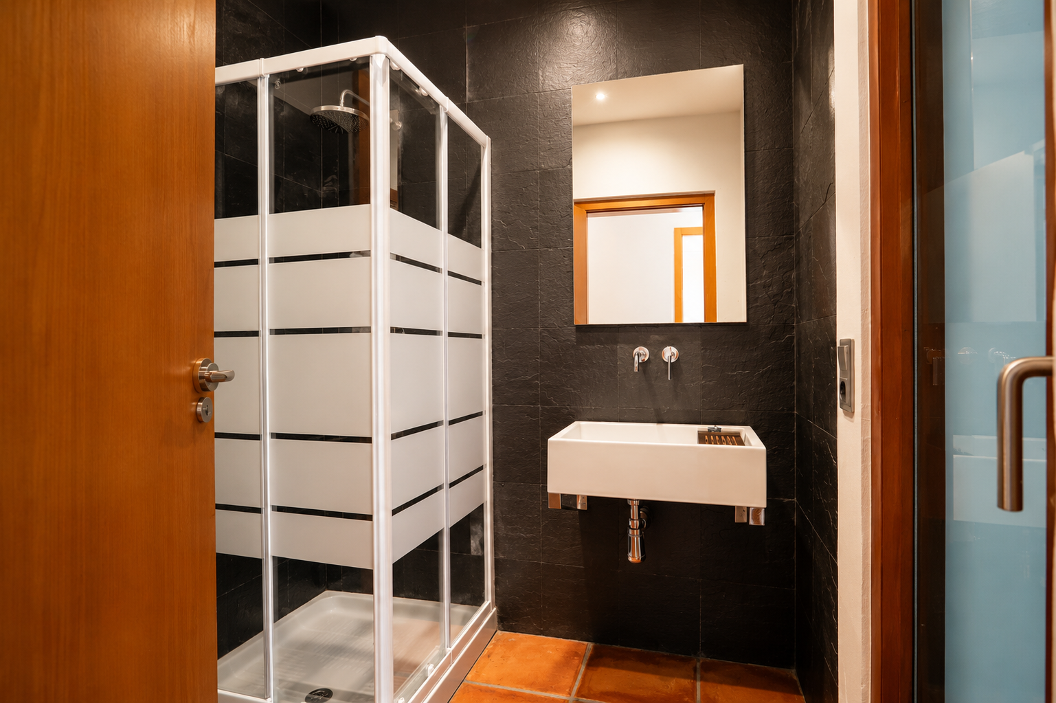 Badkamer met douchecabine met matte strepen, rechthoekige wandwastafel en donkere getextureerde wandtegels. De vloer is afgewerkt met terracotta tegels.