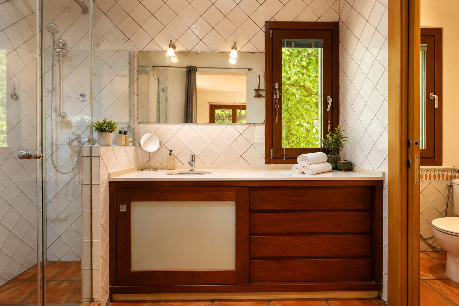 Badkamer met glazen douche, donkerhouten wastafelmeubel en wit blad. Een raam biedt uitzicht op groen, aangevuld met ruitvormige witte tegels en ambachtelijke terracotta vloeren.
