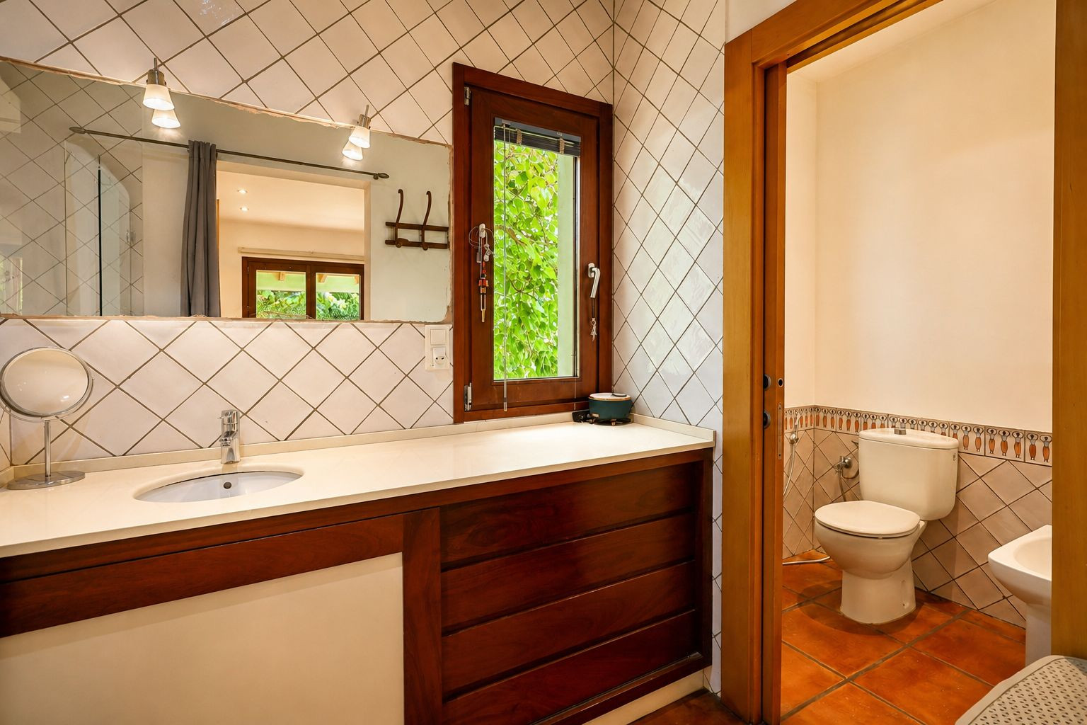 Badkamer met wastafel op licht blad en donkerhouten meubel. De grote spiegel met wandlampen en het houten raam bieden uitzicht op weelderig groen buiten.