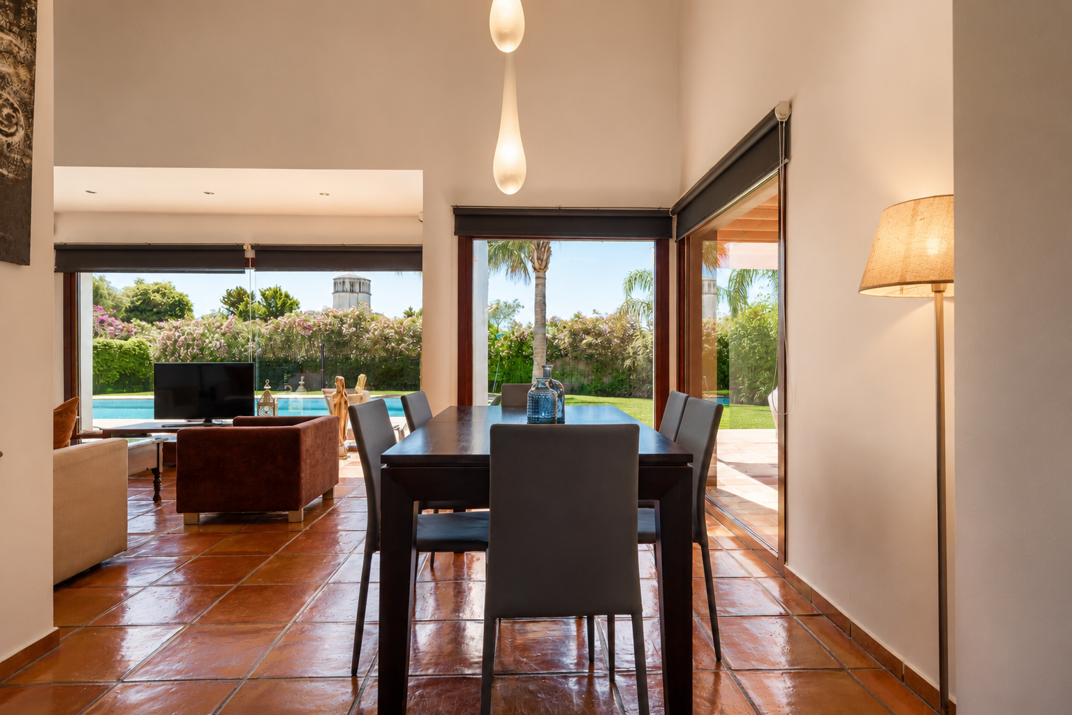 Eetruimte met donkere houten tafel en terracotta vloer, verlicht door natuurlijk licht via grote glazen deuren. Biedt direct uitzicht op de weelderige tuin, het zwembad en een verre toren.