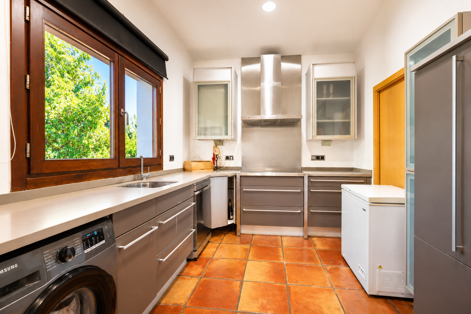 Functionele keuken met geïntegreerde wasmachine, ambachtelijke terracotta vloer en een groot houten raam met tuinzicht.