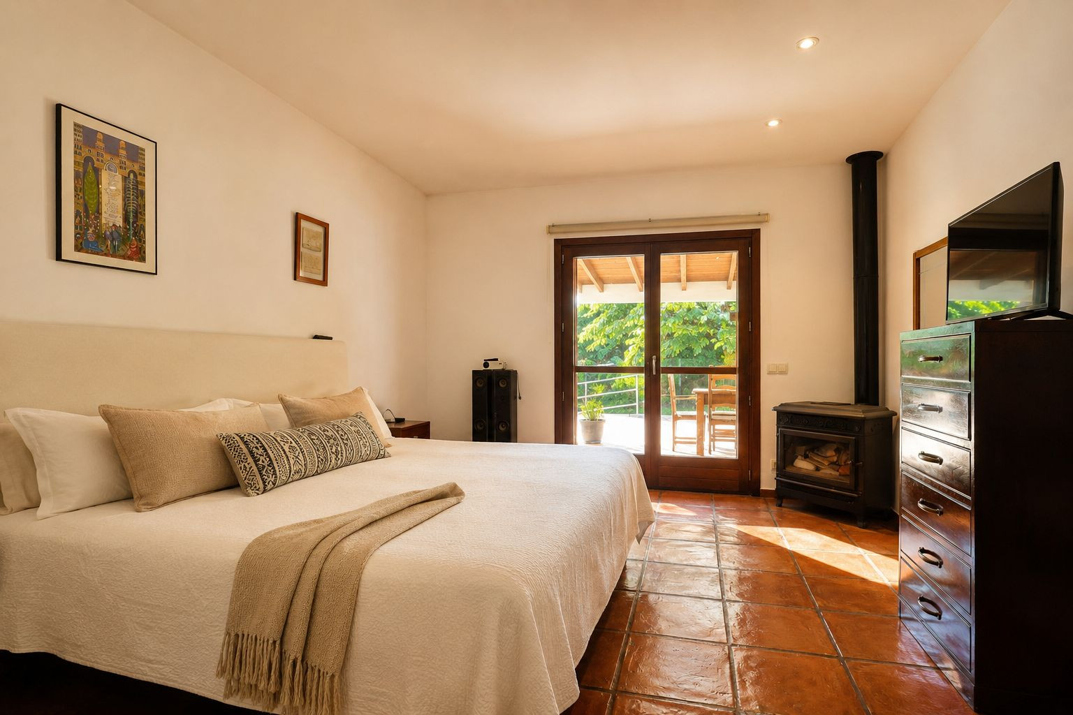 Lichte slaapkamer met een groot bed, traditionele terracotta vloertegels en een sfeervolle houtkachel. Dubbele glazen deuren bieden directe toegang tot een groen buitenterras.