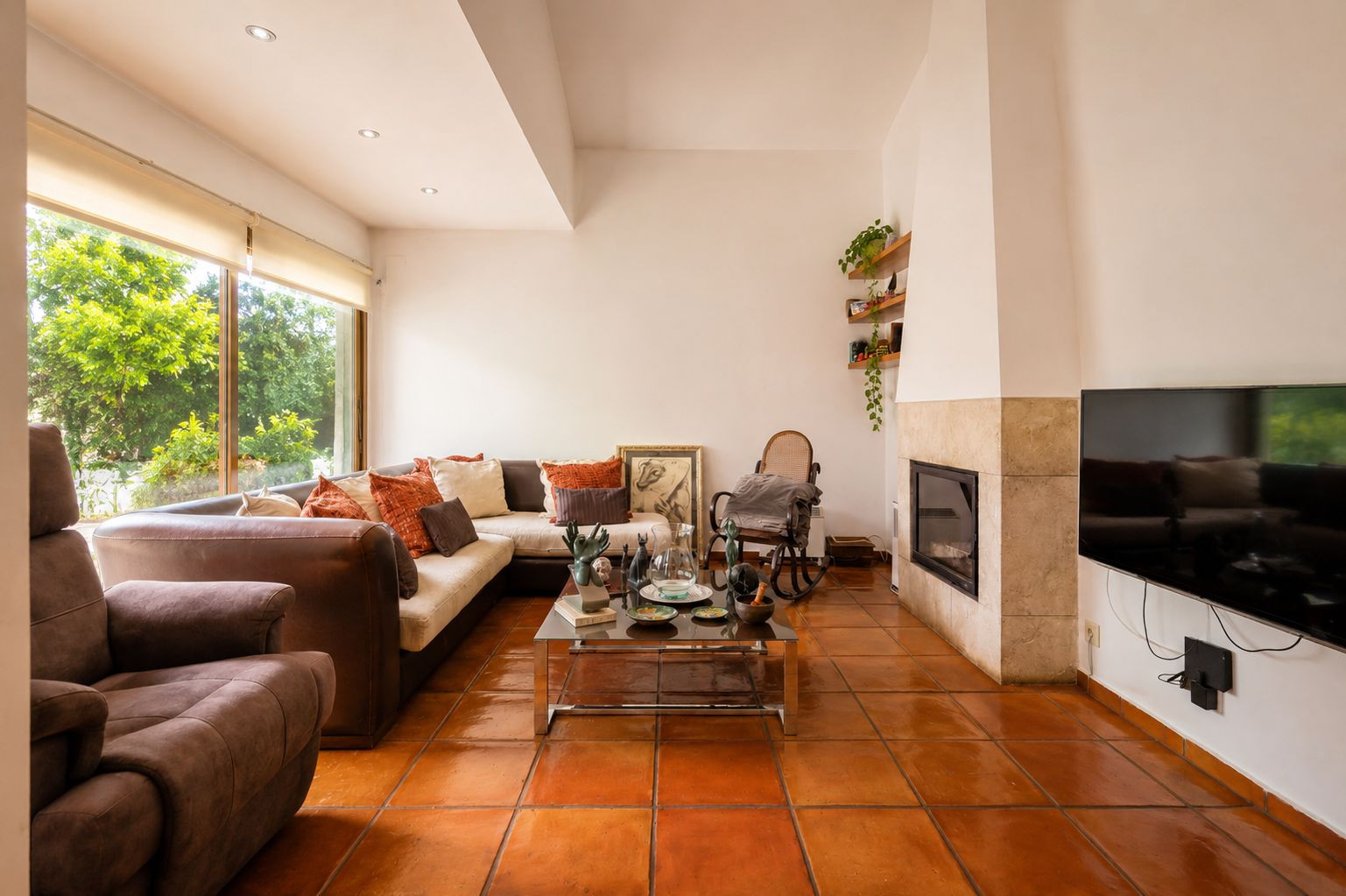 Lichte woonkamer met traditionele terracotta vloeren, een stenen open haard en grote ramen met uitzicht op weelderig groen.
