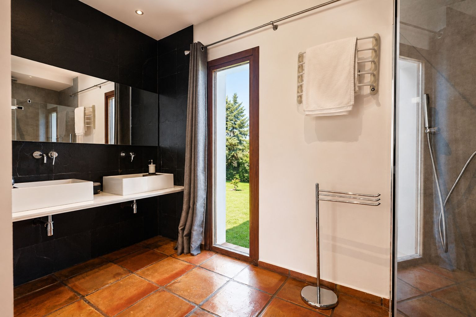 Ruime badkamer met terracotta vloertegels, dubbele witte rechthoekige wastafel op een zwarte tegelwand. Groot raam met uitzicht op de tuin en een glazen douchecabine.