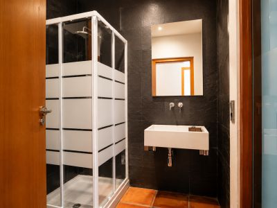 Badkamer met douchecabine met matte strepen, rechthoekige wandwastafel en donkere getextureerde wandtegels. De vloer is afgewerkt met terracotta tegels.