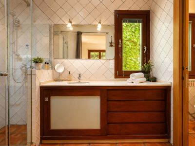 Badkamer met glazen douche, donkerhouten wastafelmeubel en wit blad. Een raam biedt uitzicht op groen, aangevuld met ruitvormige witte tegels en ambachtelijke terracotta vloeren.