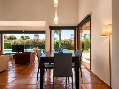 Eetruimte met donkere houten tafel en terracotta vloer, verlicht door natuurlijk licht via grote glazen deuren. Biedt direct uitzicht op de weelderige tuin, het zwembad en een verre toren.