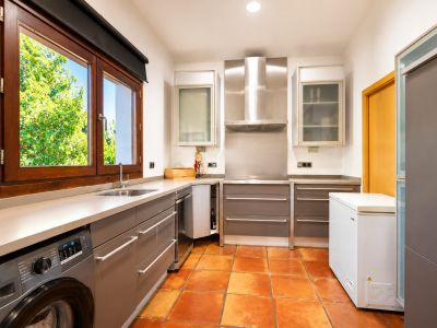 Functionele keuken met geïntegreerde wasmachine, ambachtelijke terracotta vloer en een groot houten raam met tuinzicht.