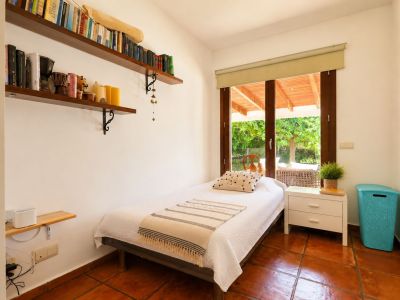 Individuele slaapkamer met terracotta tegelvloer, witte muren en directe toegang tot een overdekt terras via een grote houten glazen deur. Voorzien van wandplanken met boeken.
