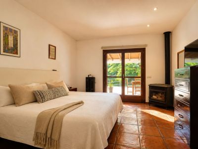 Lichte slaapkamer met een groot bed, traditionele terracotta vloertegels en een sfeervolle houtkachel. Dubbele glazen deuren bieden directe toegang tot een groen buitenterras.