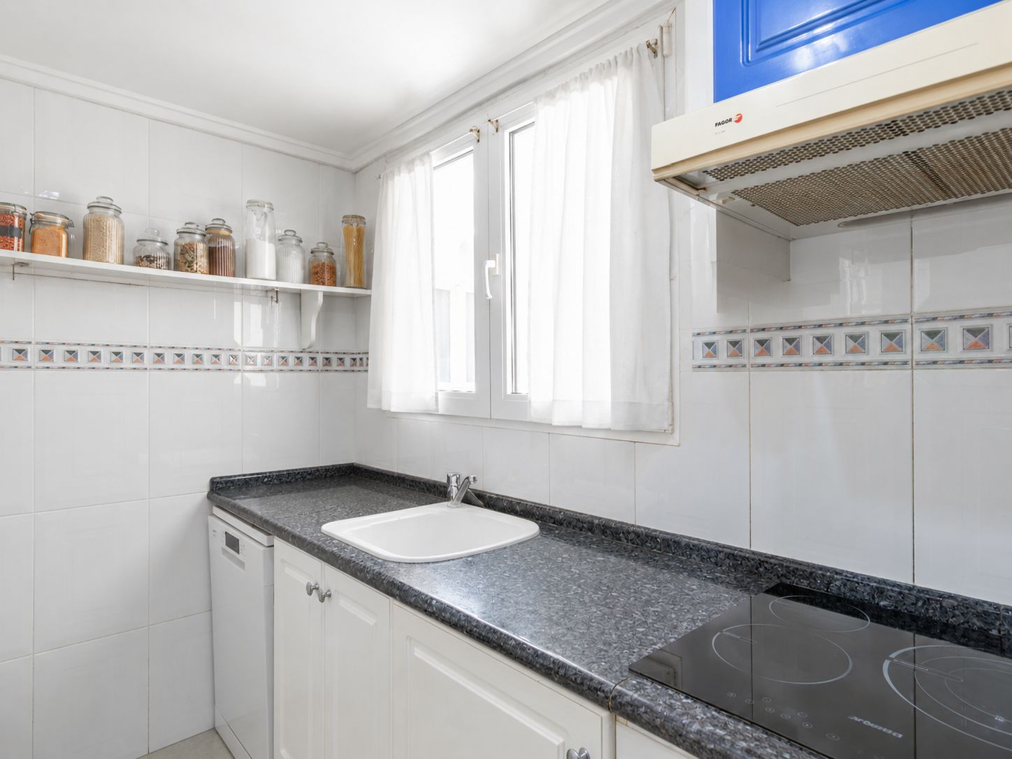 Aparte keuken met donker granieten aanrechtblad, witte spoelbak en inductiekookplaat. Witte tegelwanden met decoratieve rand en raam voor natuurlijk licht.