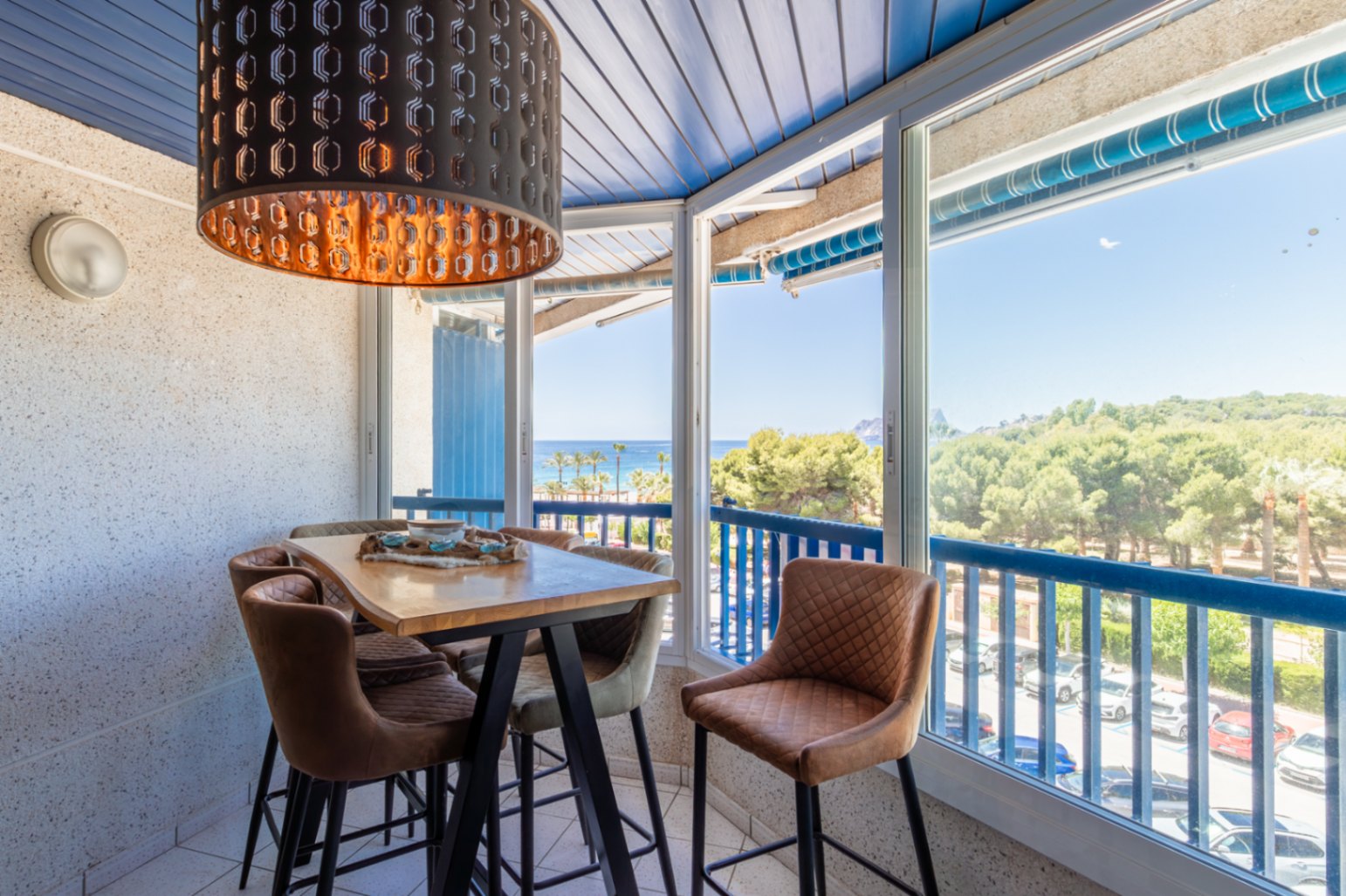Beglaasde eethoek met hoge houten tafel, gestoffeerde barkrukken en opvallende zwarte hanglamp. Panoramisch uitzicht op zee, strand en dennenbos door grote ramen.